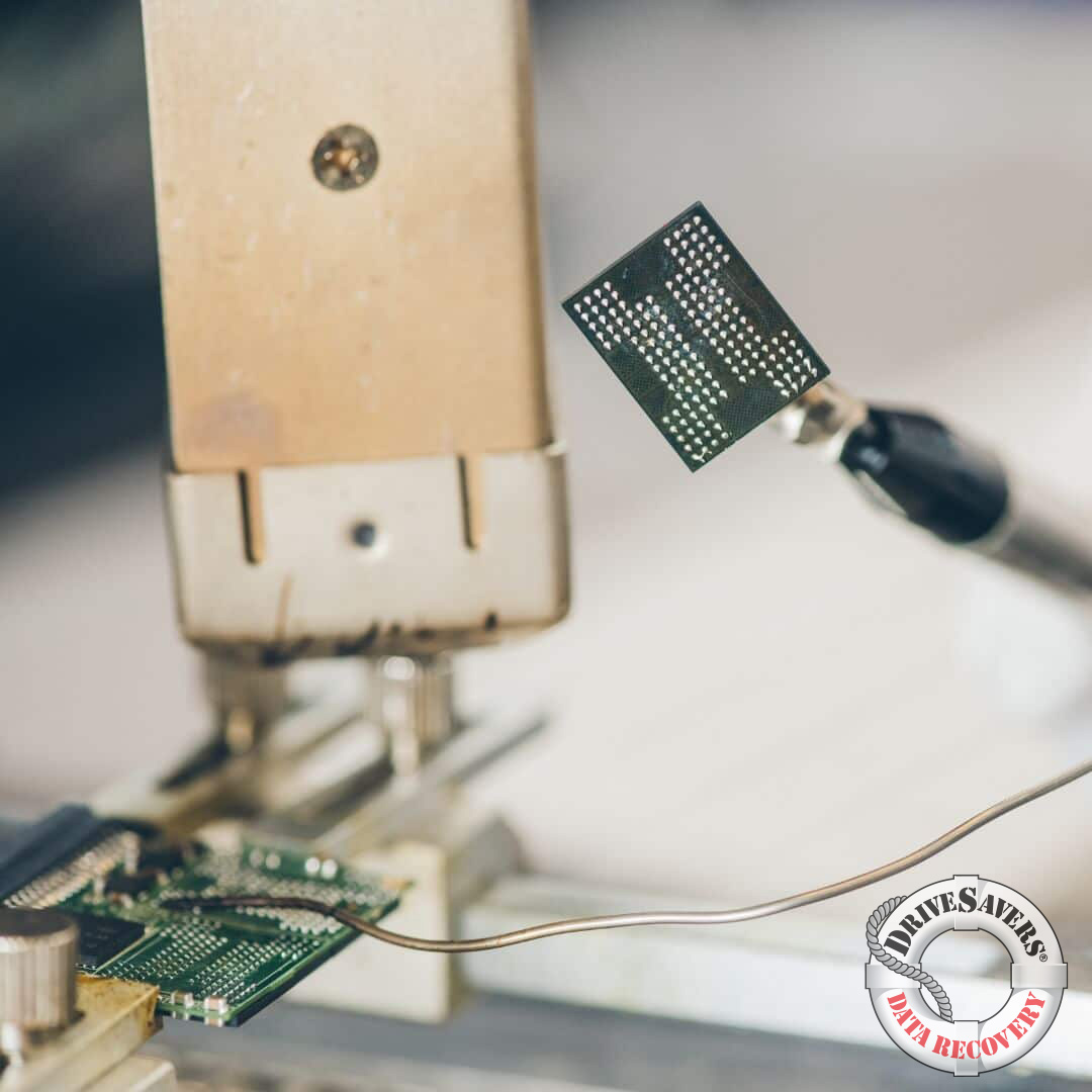 A close-up of microchip repair tools handling electronic components is shown. The background features a blurred workbench setting, with the "DriveSavers Data Recovery" logo visible in the corner.