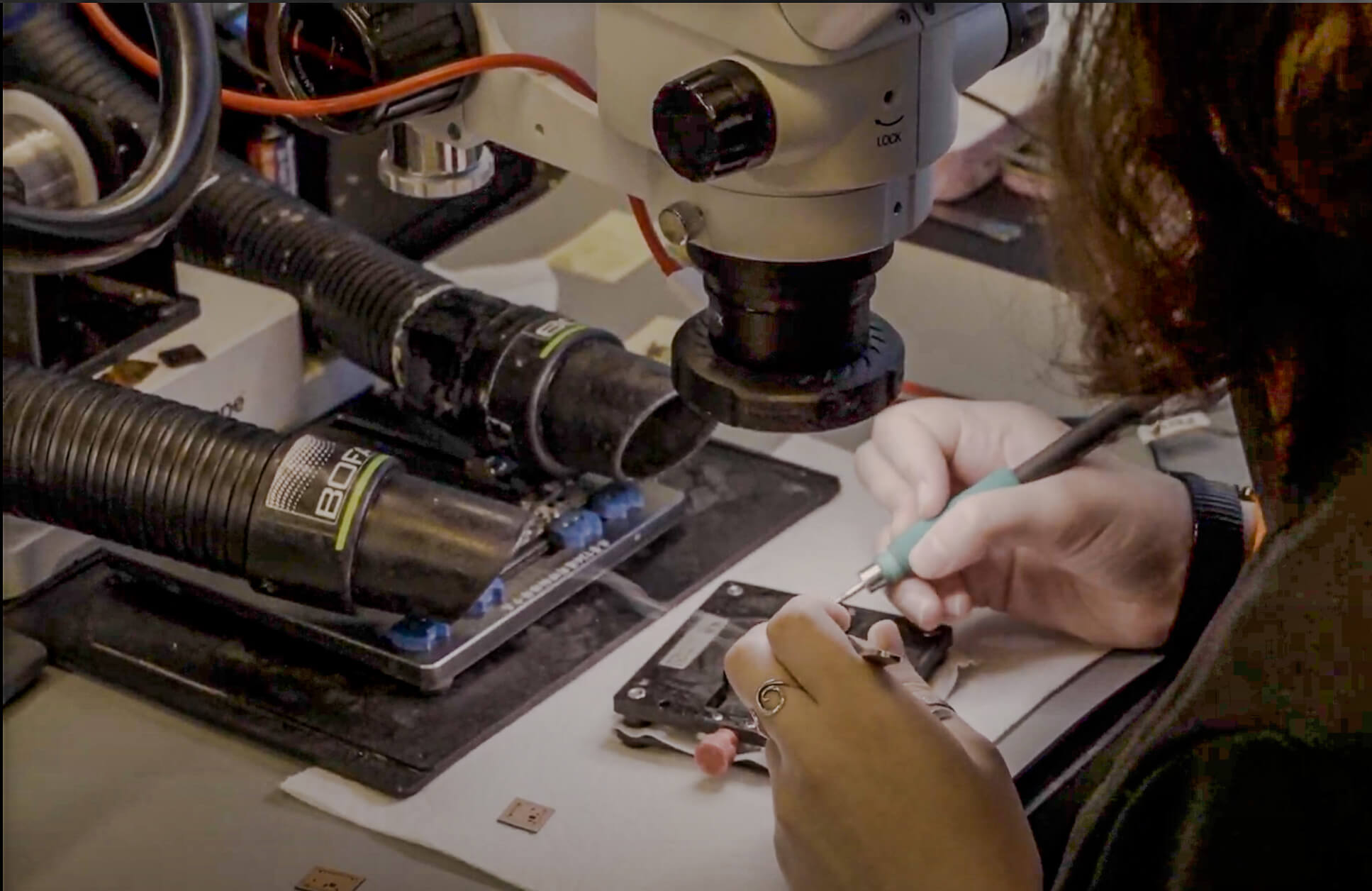 Person using a microscope and tools to work on small electronic components at a workstation, with visible ventilation hoses and a circuit board in focus.