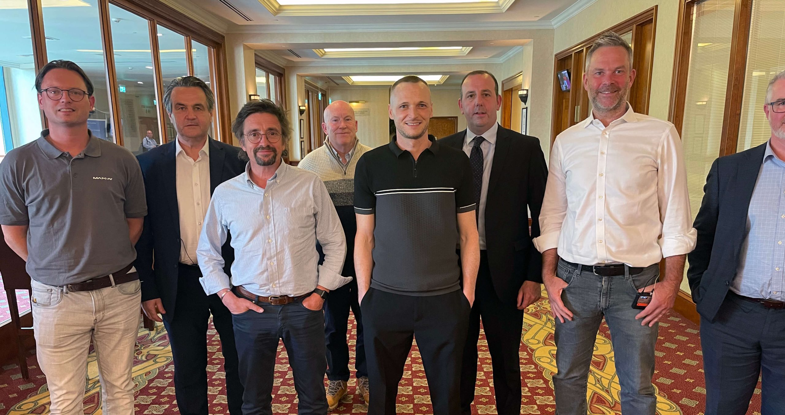 A group of eight men stand indoors, posing for a photo. Some are in casual attire, while others wear suits. The room has large windows and a patterned carpet. Amidst the chatter of the event, one jokes about how his external hard drive keeps disconnecting at work.