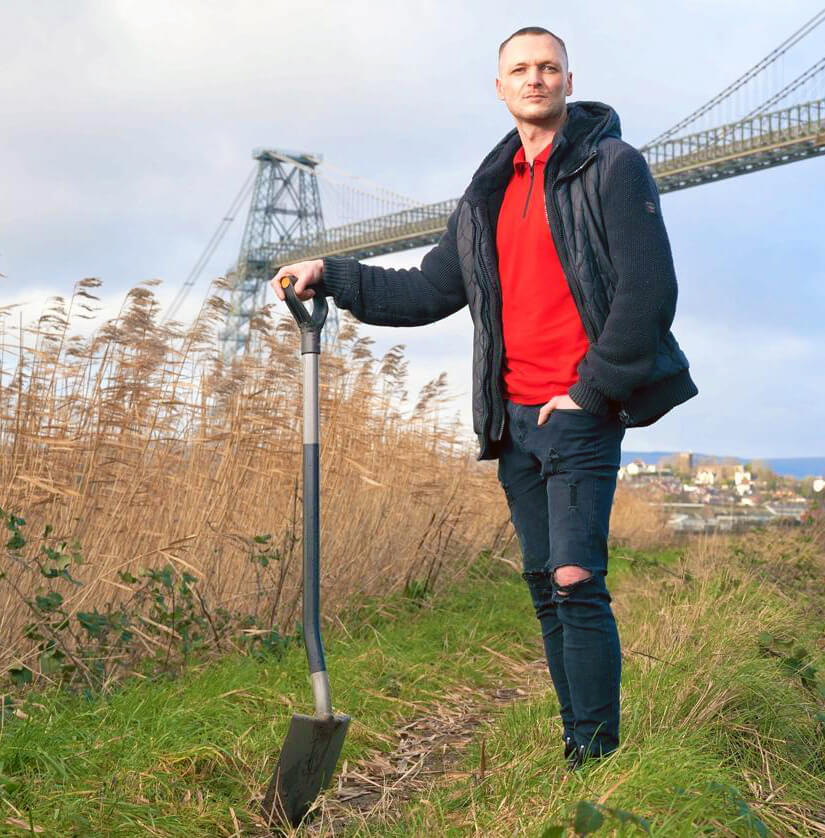 A person stands on a grassy path holding a shovel, their red shirt and dark jacket contrasting with the overcast sky. Tall grasses surround them as if offering a natural embrace. In the background, a large bridge looms, much like an external hard drive keeps disconnecting from its digital world.